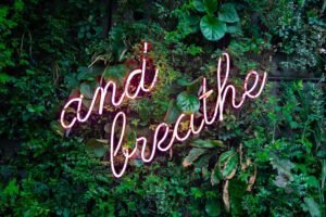 Neon sign saying 'and breathe' amid lush green leaves