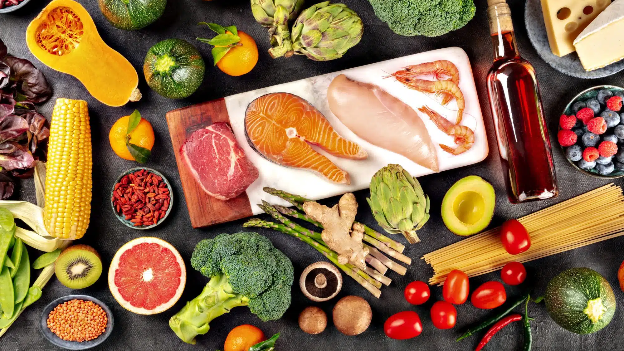 Assorted fresh food ingredients including salmon, shrimp, broccoli, and more on a dark background