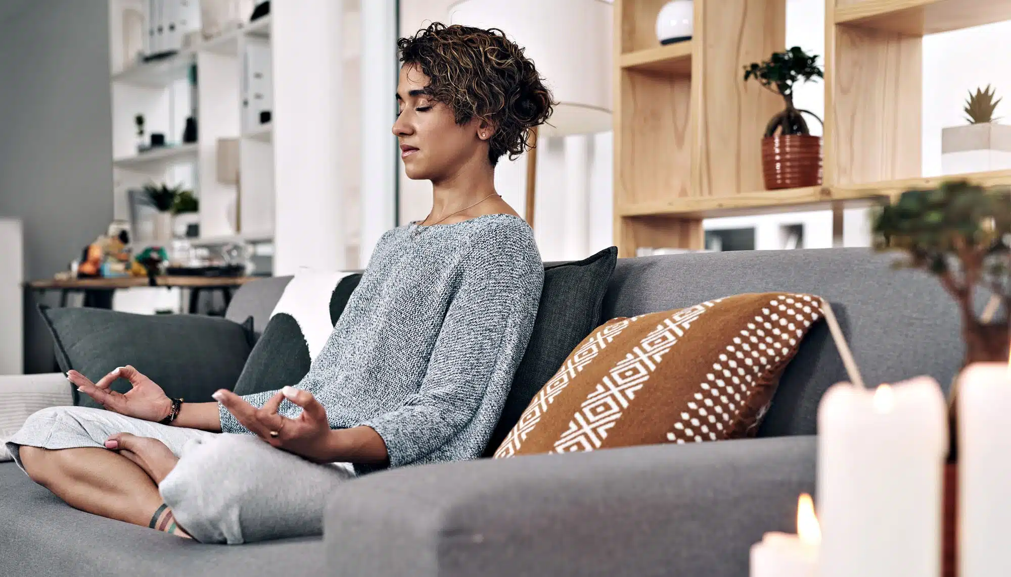 Young woman meditating on sofa, surrounded by candles and plants