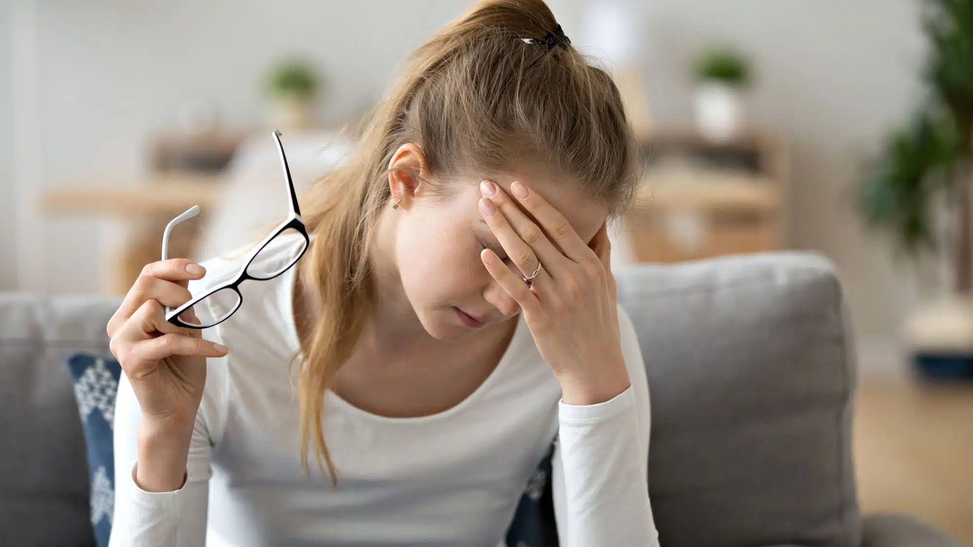 Woman with headache holding glasses, sitting on couch in living room