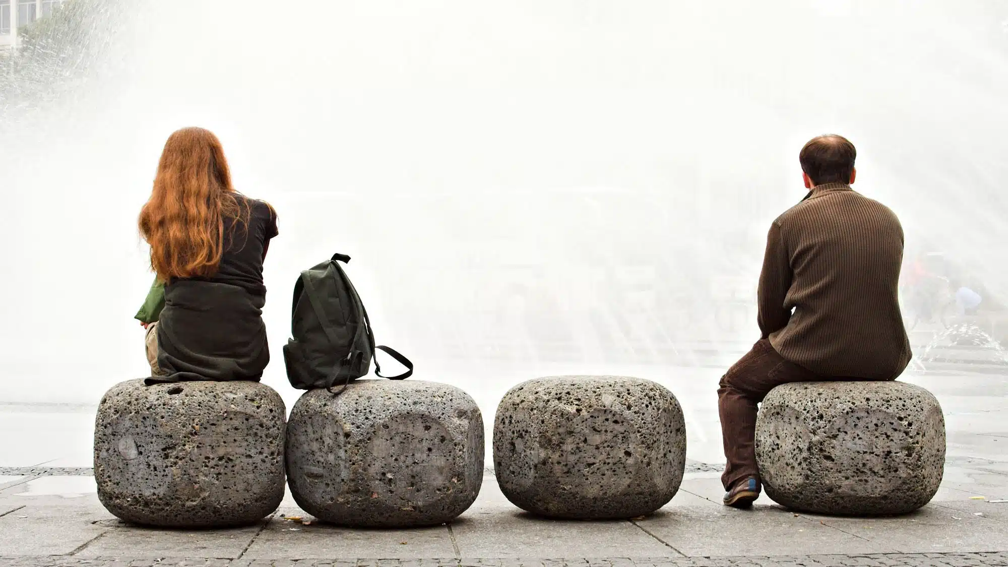 Two people sitting back to back on stone benches near a water fountain